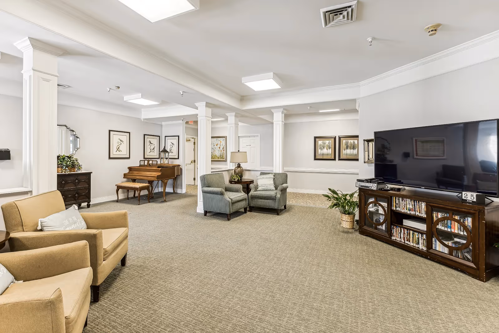 A spacious living room area with beige carpet and white walls. The room features two beige armchairs with pillows on the left, two green armchairs with a small table and lamp in the center, and a large flat-screen TV on a wooden cabinet filled with DVDs on the right. There is a wooden piano with a matching bench against the far wall, decorated with framed artwork and a mirror. The ceiling has recessed lighting and white columns separate the space.
