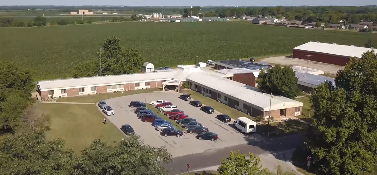 Aerial view of a single-story L-shaped building surrounded by trees and fields, with a parking lot filled with cars in front. The building appears to be a healthcare or residential facility.