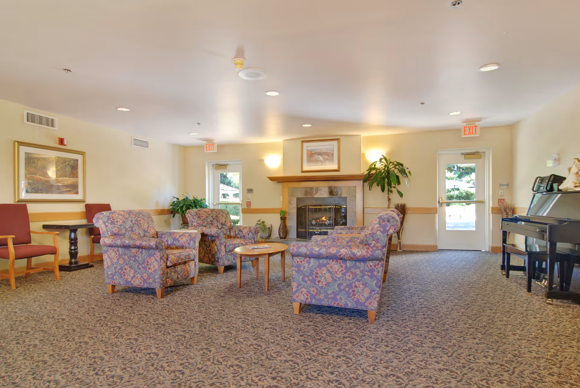 Spacious senior living lounge with patterned armchairs arranged around a coffee table facing a fireplace, with a piano and exit doors in the background.