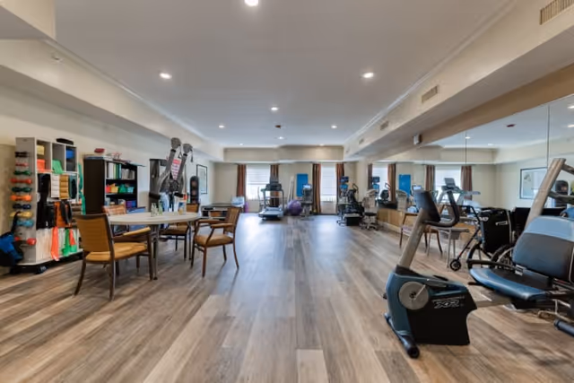 A spacious fitness room in a senior living facility with exercise equipment including stationary bikes, treadmills, and weights. There are tables and chairs on the left side, shelves with exercise accessories, and large mirrors on the right wall. The room has wood flooring and recessed ceiling lights.