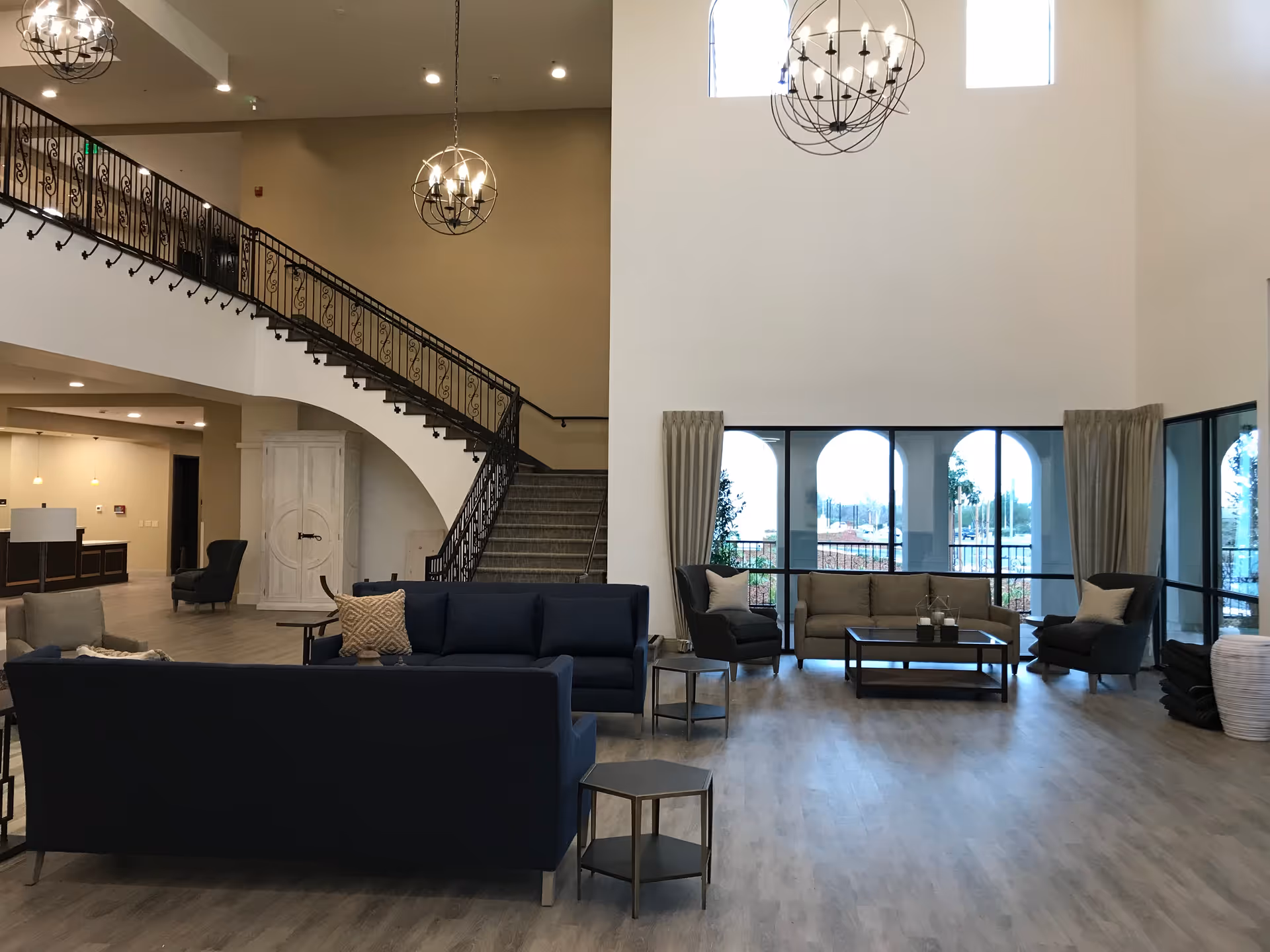 Spacious high-ceilinged common lounge with sofas and armchairs, large windows, hanging chandeliers and a curved staircase.