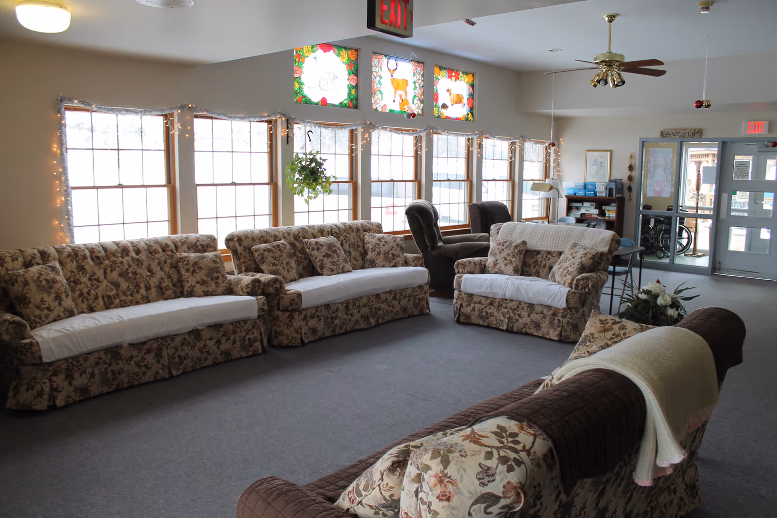 Bright communal living room with floral sofas and armchairs arranged in front of large windows with stained-glass transoms and an exit door in the background.