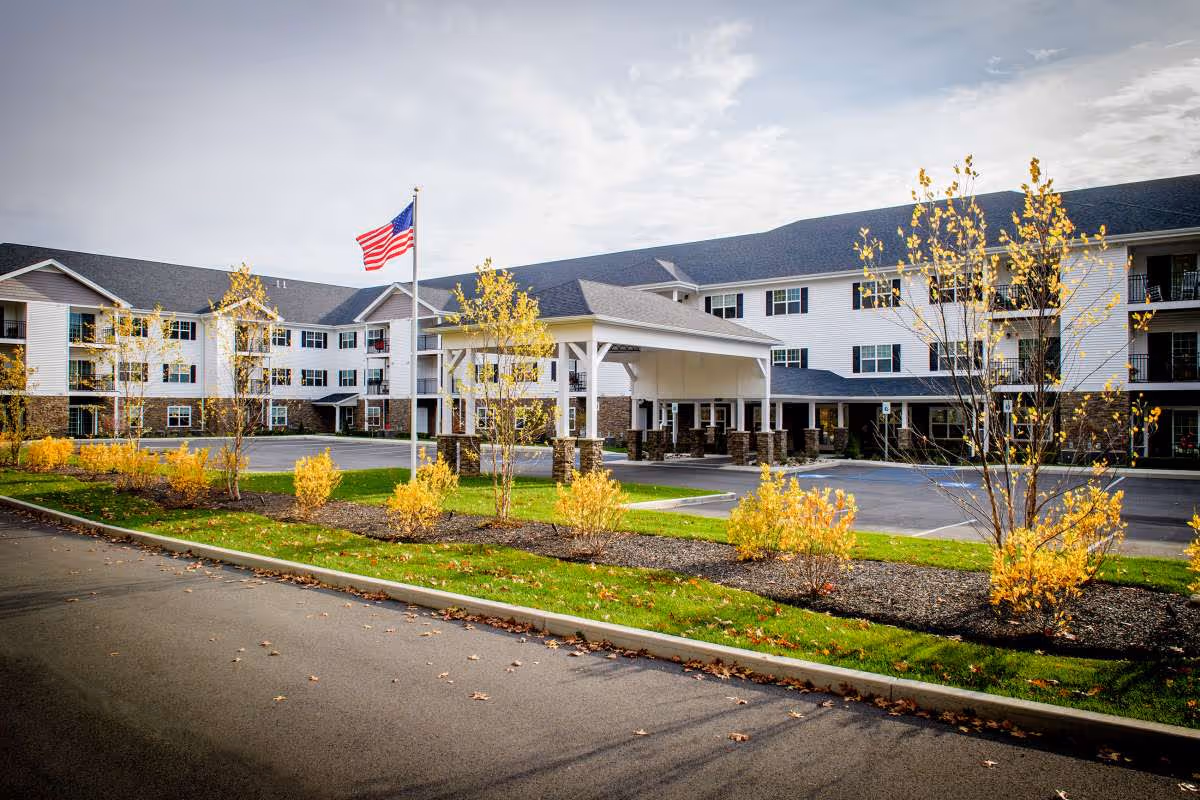 Exterior view of a large senior living facility building with white siding and stone accents, a covered entrance, an American flag on a flagpole, and landscaped grounds with small trees and shrubs showing fall colors.