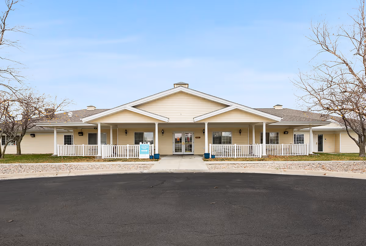 Front exterior view of a single-story senior living facility building with beige siding, a covered entrance with white railings, and leafless trees on either side under a clear sky.