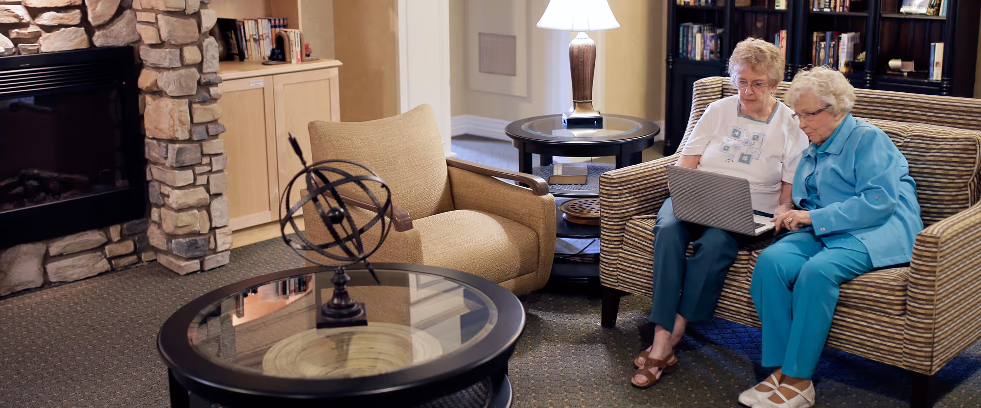 Two elderly women sitting on a striped sofa in a cozy living room area, looking at a laptop together. The room features a stone fireplace, a beige armchair, a round glass coffee table with a decorative globe, a side table with a lamp, and bookshelves in the background.