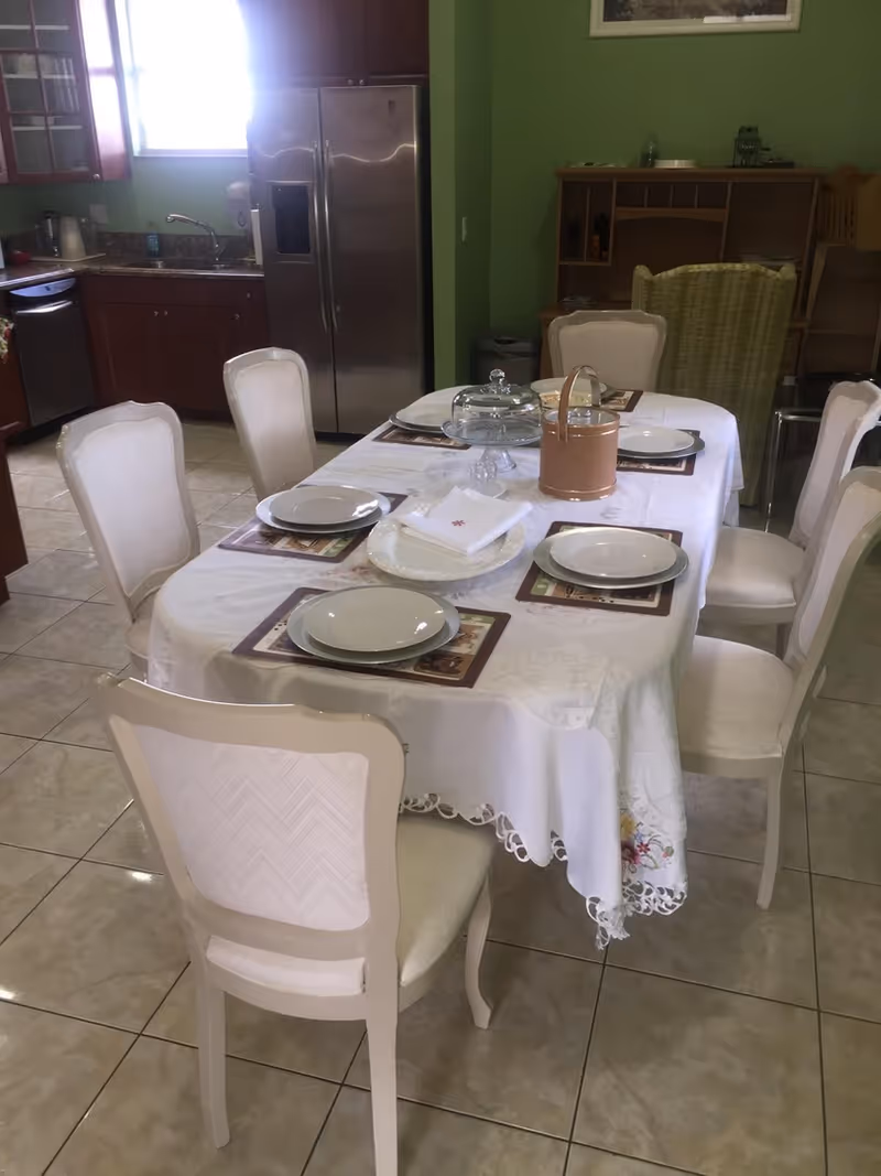 A dining area with a rectangular table covered with a white tablecloth and set with six place settings, each with a plate and placemat. The table also has a glass cake stand with a cover and a small basket. Surrounding the table are six white cushioned chairs. In the background, there is a kitchen with wooden cabinets, a stainless steel refrigerator, a dishwasher, and a sink under a window. The walls are painted green and the floor is tiled.