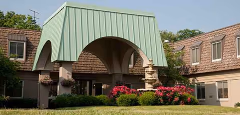 Arched green roof canopy over entrance of a senior living facility with flowering shrubs.