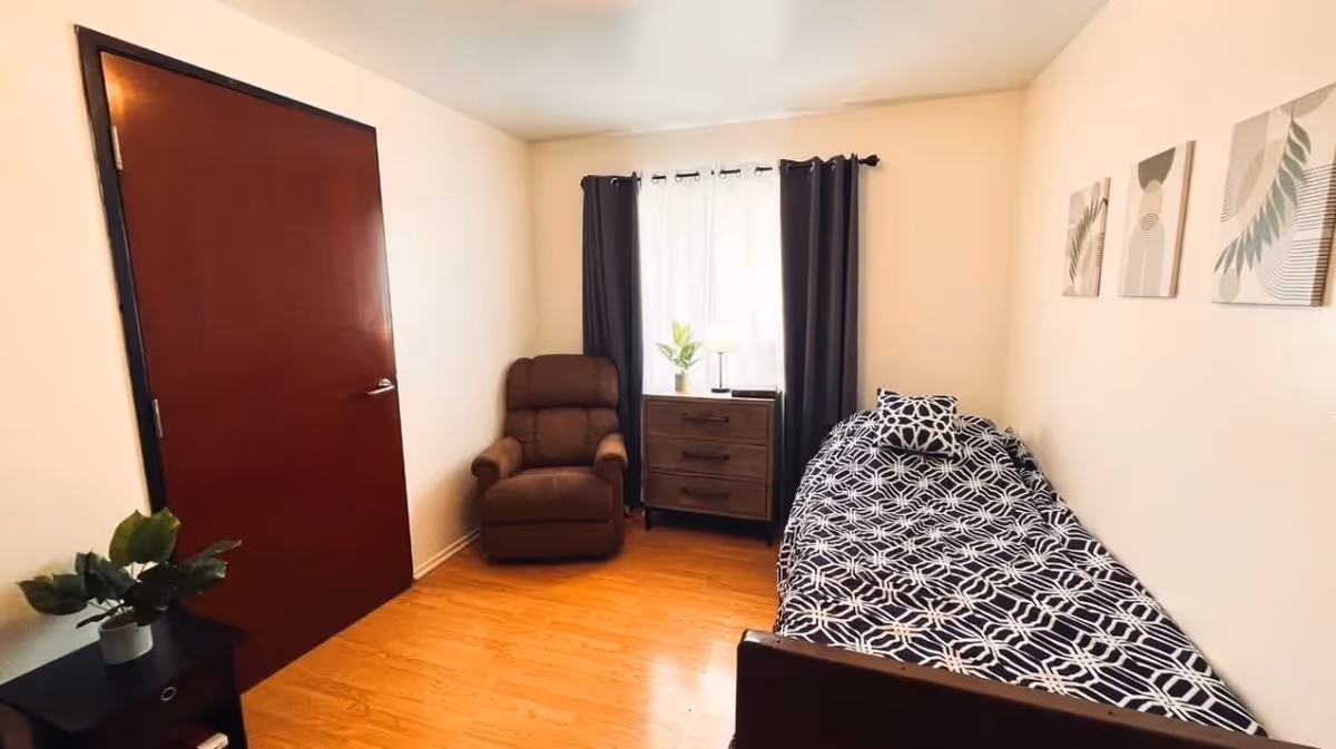 A small bedroom with a single bed covered in a black and white geometric patterned bedspread and matching pillow. There is a brown recliner chair next to a window with dark curtains and a sheer white curtain. A wooden dresser with three drawers is placed under the window, holding a small lamp and a potted plant. The walls are light-colored with three abstract art pieces hanging above the bed. The floor is wooden, and a closed dark red door is visible on the left side of the room.