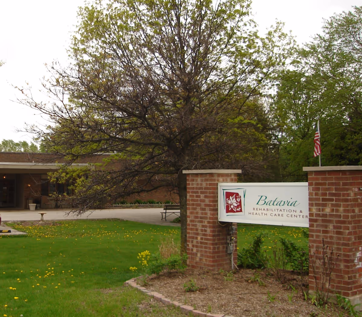Exterior view of Batavia Rehabilitation & Health Care Center showing a brick sign with the facility's name, a large tree, green lawn with some yellow flowers, and part of the building entrance in the background.