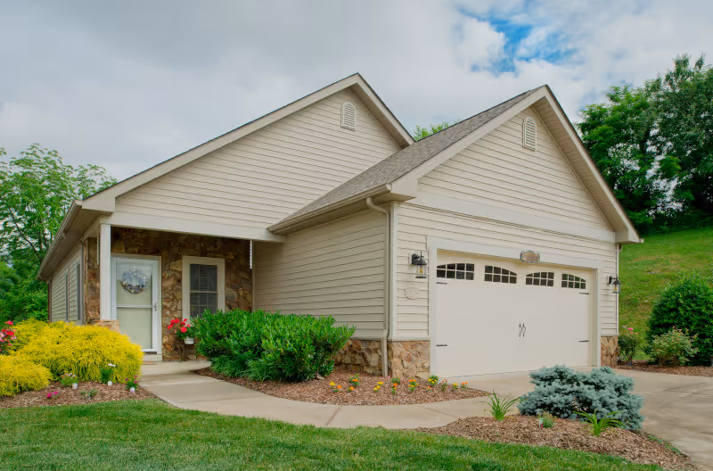 Exterior view of a single-story residential building with beige siding and stone accents. The building has a two-car garage with white doors and a small covered entrance with a white door and window. The surrounding area features a well-maintained lawn, shrubs, and small trees under a partly cloudy sky.