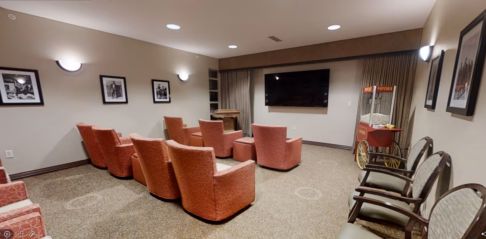 A cozy media room with several orange patterned armchairs arranged in rows facing a large flat-screen TV mounted on the wall. There is a vintage-style popcorn machine on the right side of the room and framed black-and-white photographs hanging on the beige walls. The room is carpeted and softly lit by wall sconces and recessed ceiling lights.