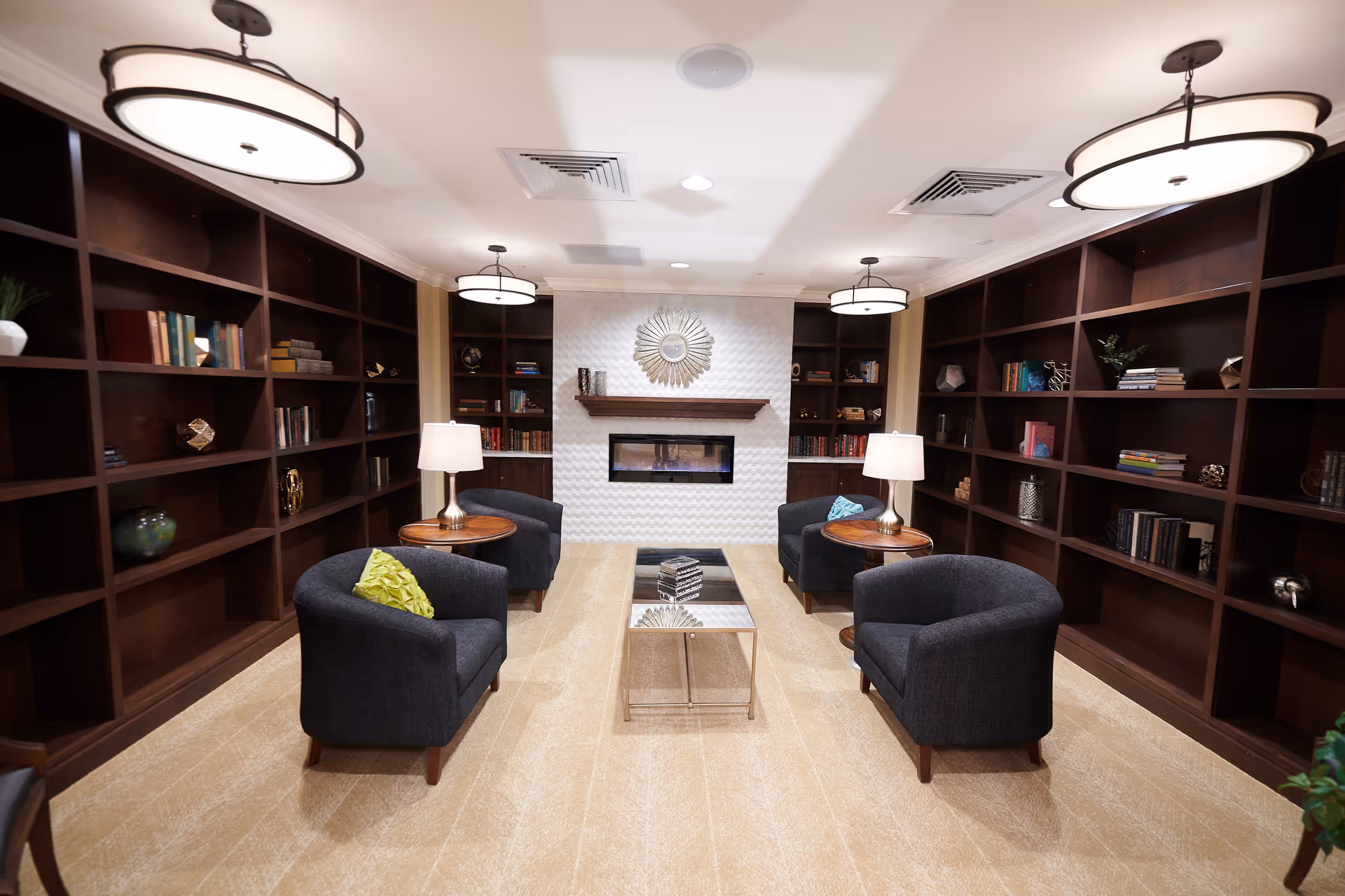 A cozy senior living common area with four dark upholstered armchairs arranged around a glass coffee table. The room features built-in dark wood bookshelves filled with books and decorative items on both sides. At the far end, there is a modern electric fireplace with a decorative sunburst mirror above it. Two round wooden side tables with lamps are placed between the chairs. The ceiling has three round pendant lights, and the floor is carpeted in a light beige color.