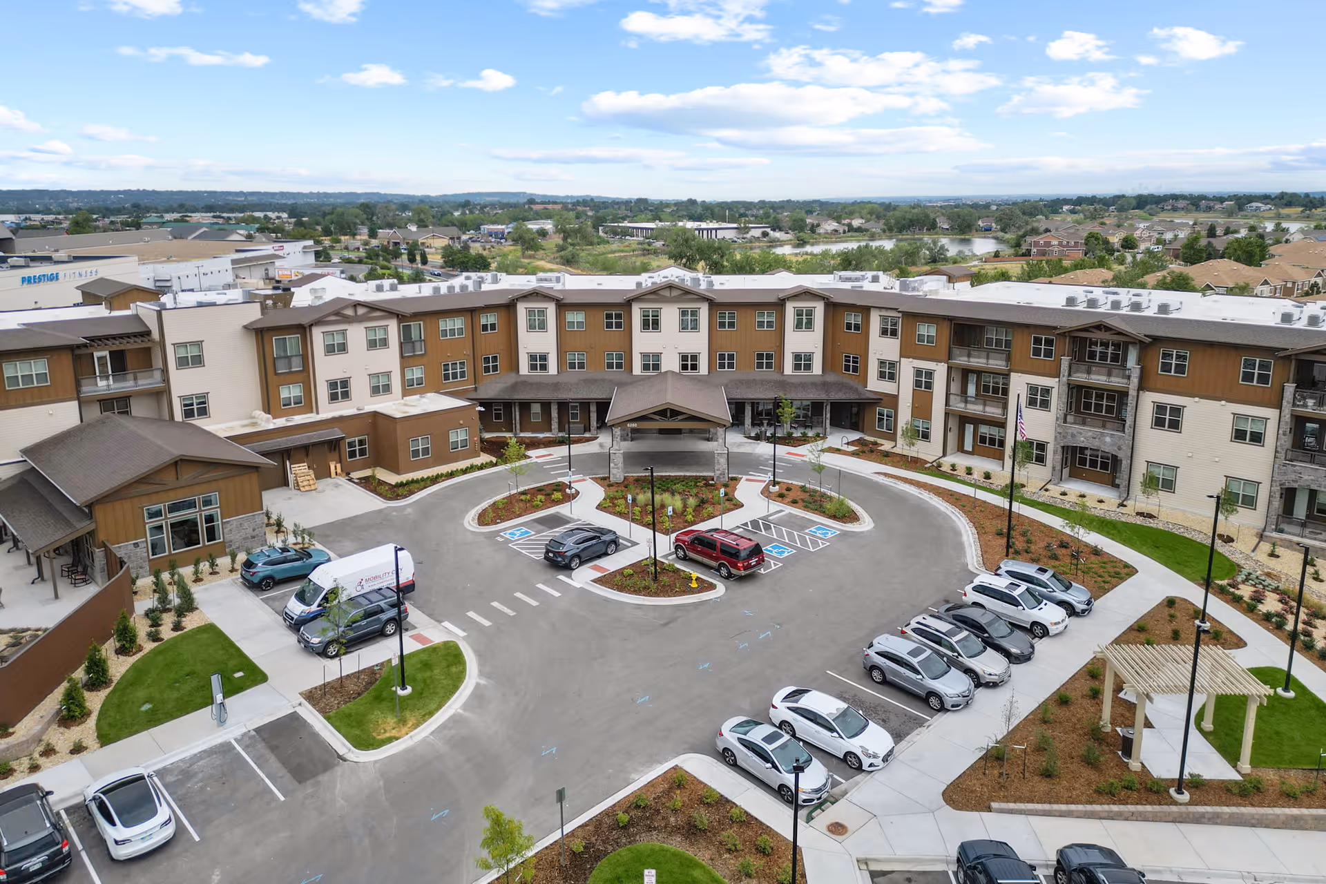 Aerial view of Amira Choice Arvada senior living facility showing a large U-shaped building with multiple floors, a covered entrance, parking spaces including handicapped spots, landscaped areas with grass and small trees, and a clear sky with scattered clouds in the background.