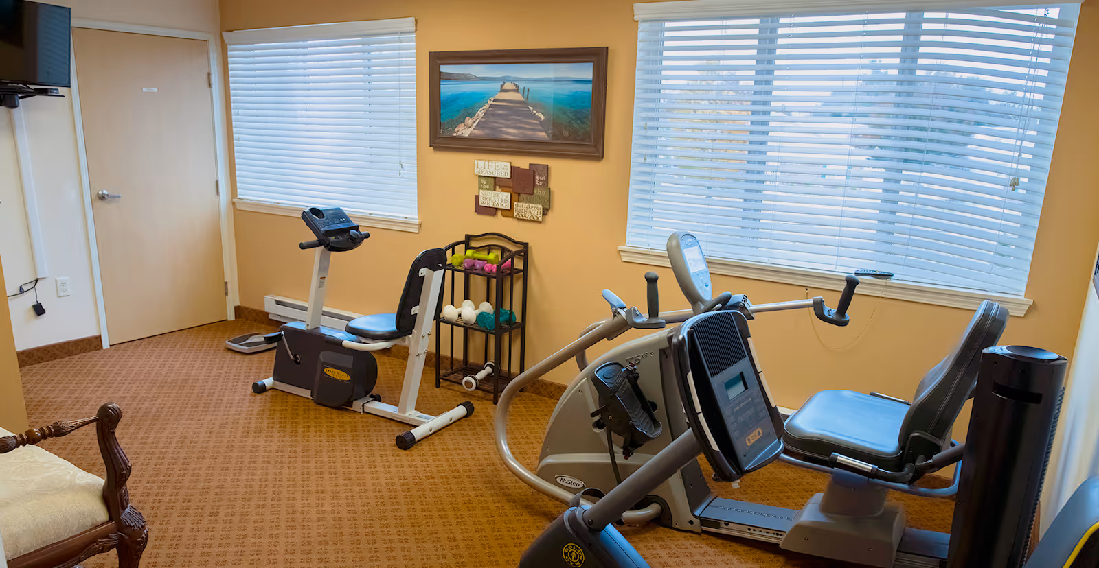 Small exercise room with two recumbent bikes, a rack of light dumbbells, and windows with blinds.