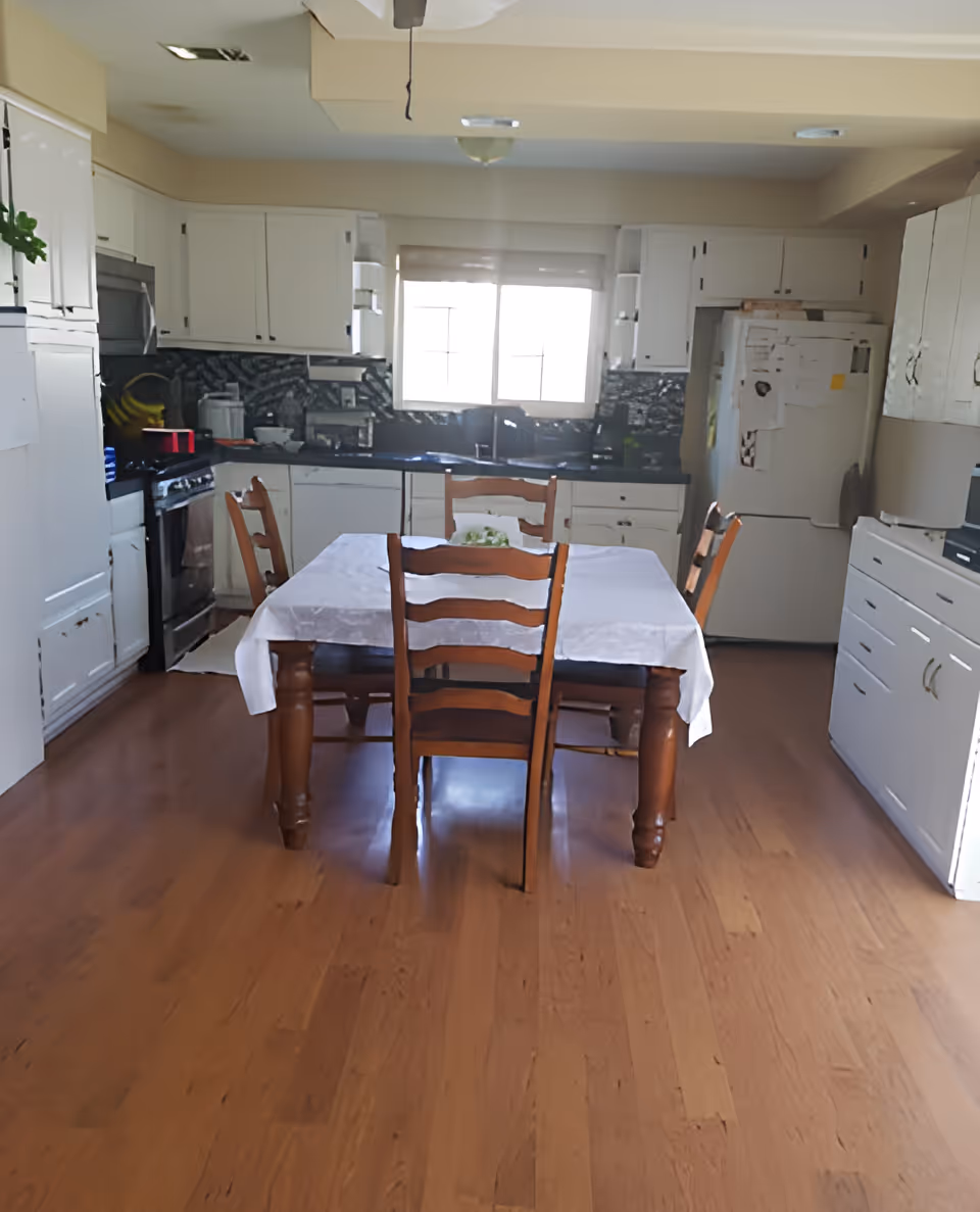 A kitchen with wooden flooring, white cabinets, a refrigerator covered with papers, a stove, and a dining table with four wooden chairs and a white tablecloth. A window above the sink lets in natural light.