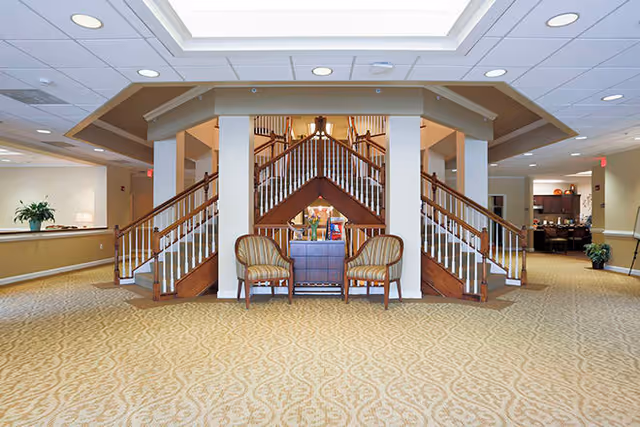 Spacious interior area with a central wooden staircase featuring two symmetrical flights of stairs and a small seating area with two striped armchairs and a wooden table in between. The room has beige patterned carpet, recessed lighting, and a skylight above the staircase. There are hallways visible on either side with plants and furniture.