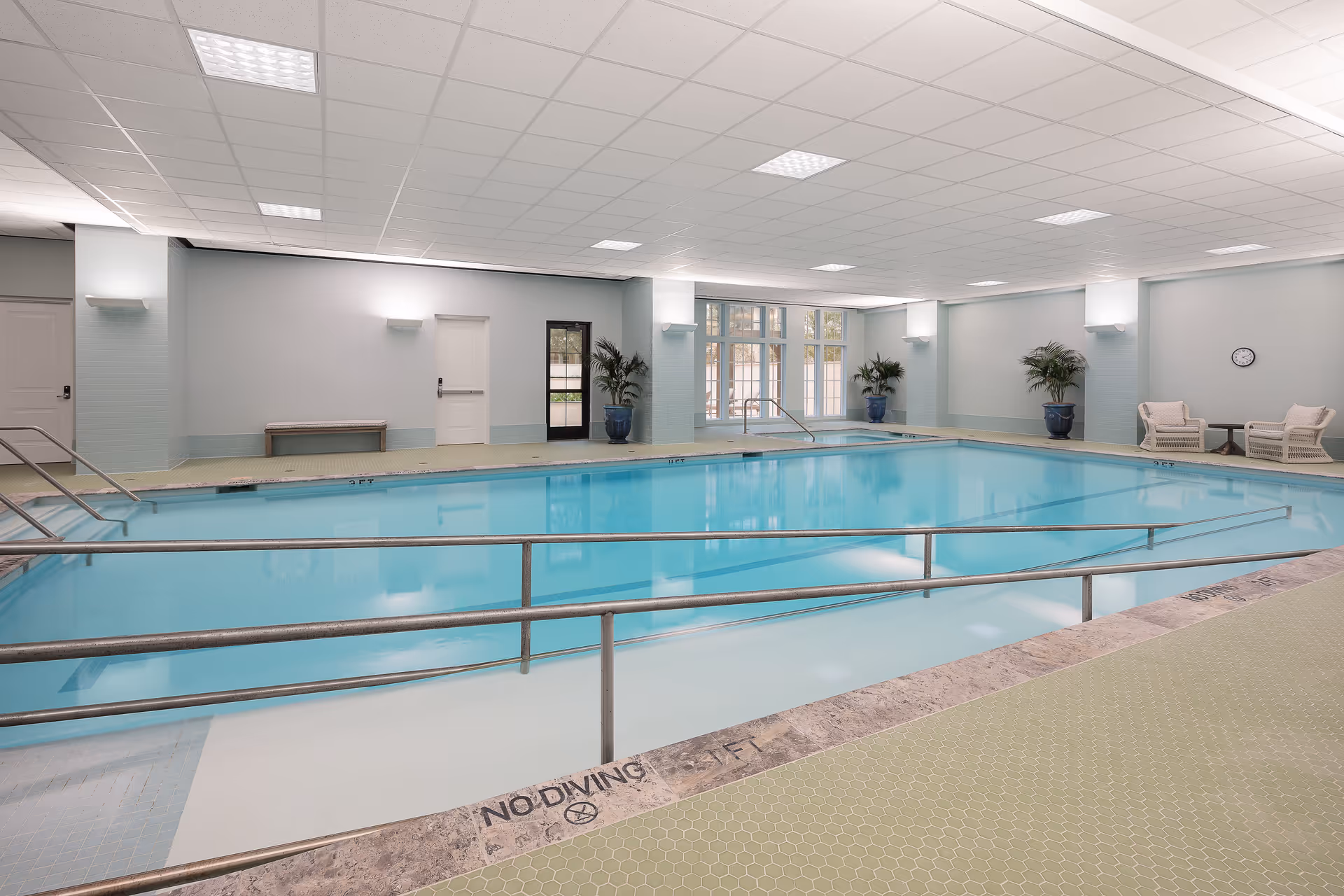 Large indoor swimming pool with handrails, lounge chairs, and potted plants in a light-colored room.
