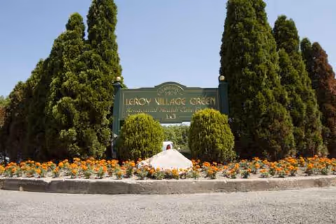 Entrance sign reads Leroy Village Green, framed by tall evergreens and flowers.