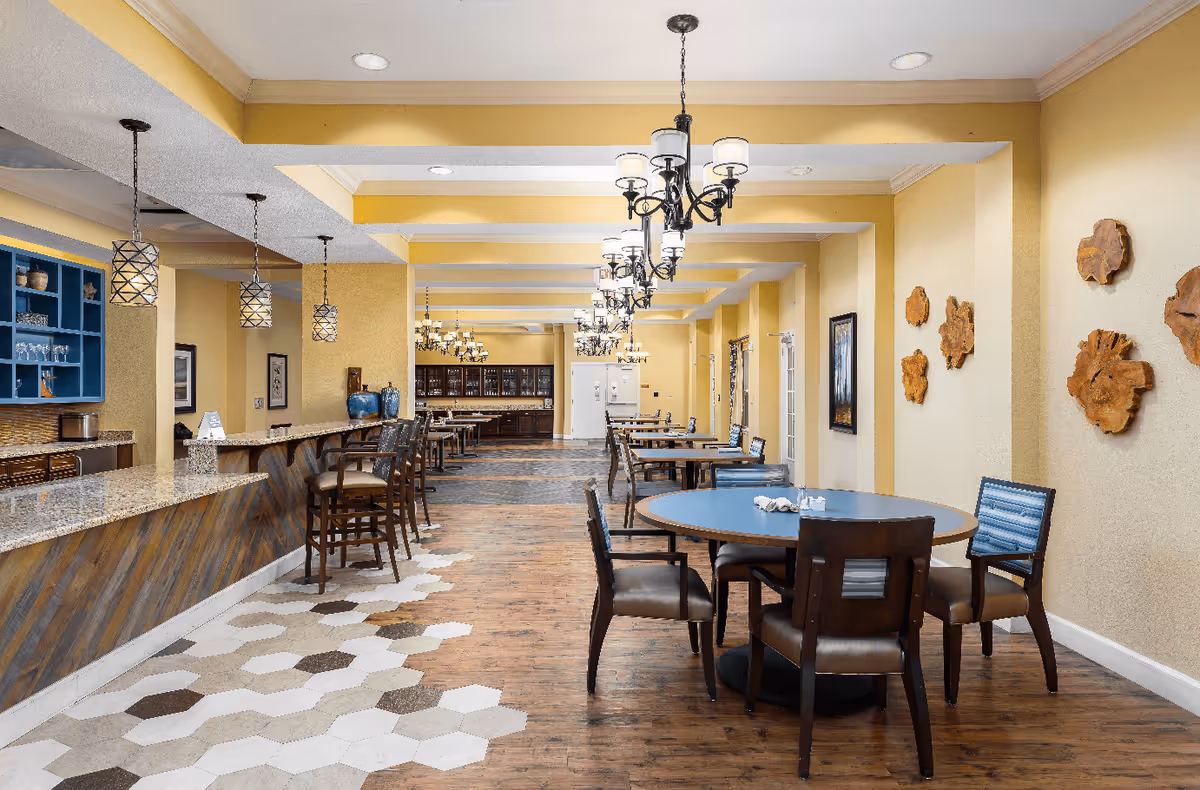 A spacious dining area with multiple round and rectangular tables surrounded by chairs. The room features yellow walls with decorative wooden wall art, a long counter with bar stools, pendant lights, and chandeliers hanging from the ceiling. The floor transitions from hexagonal tiles near the counter to wood flooring in the seating area.