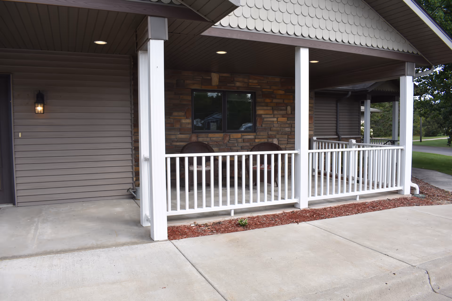 Covered porch area with white railing and two brown wicker chairs against a stone wall exterior. The porch is part of a building with brown siding and a concrete walkway in front.