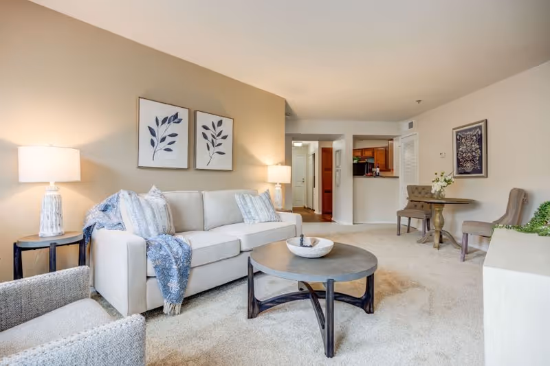 A bright and cozy living room in a retirement community featuring a beige sofa with decorative pillows and a blue throw, a round wooden coffee table with a decorative bowl, two framed botanical prints on the wall, a side table with a lamp, and a small dining area with two upholstered chairs and a round table. The room has beige carpeting and neutral-colored walls, with a view into a kitchen area in the background.