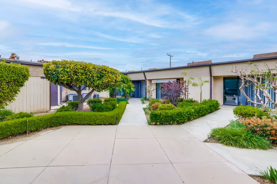 Outdoor courtyard area of Arcadia Healthcare Center featuring a paved walkway surrounded by neatly trimmed bushes, small trees, and various plants. The building exterior is visible with multiple doors and windows, under a partly cloudy sky.