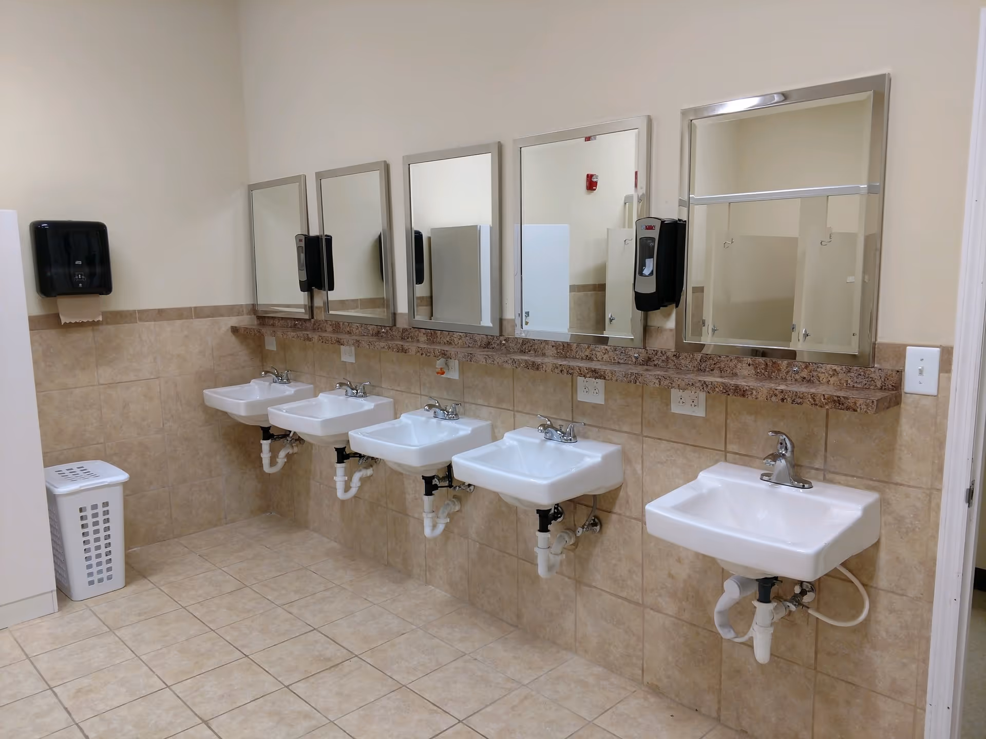 A clean restroom area with five white sinks mounted on a tiled wall. Above each sink is a rectangular mirror with a silver frame. There are two black soap dispensers mounted on the wall between the mirrors. The floor and lower half of the wall are covered with beige tiles. A white laundry basket is placed in the corner to the left.