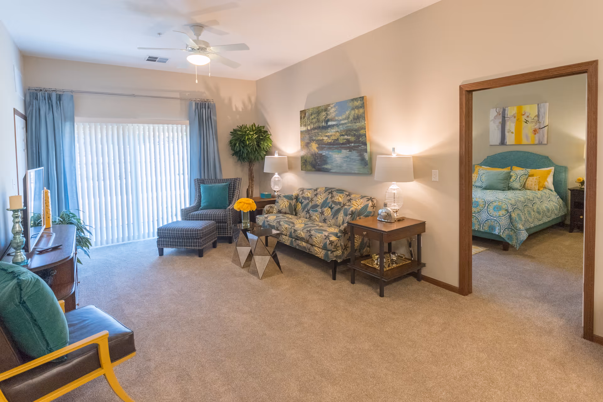 A cozy living room with beige walls and carpet, featuring a floral patterned sofa, a checkered armchair with matching ottoman, a wooden side table with lamps, and a painting on the wall. A doorway leads to a bedroom with a blue and yellow bedspread and artwork above the bed. Large vertical blinds cover a sliding glass door, and a ceiling fan with a light is mounted above.
