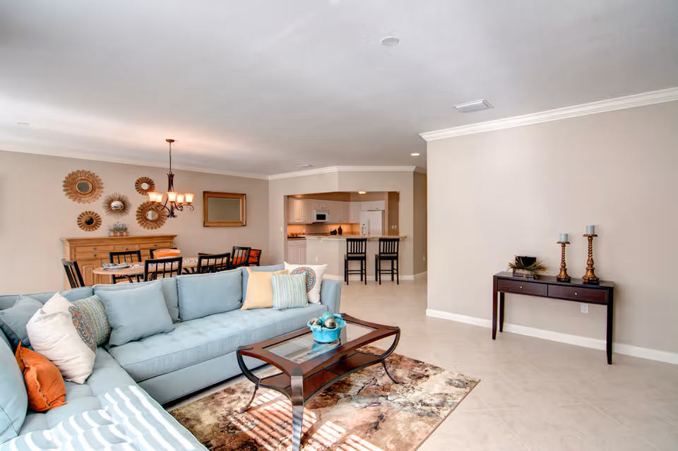 Bright and spacious living room with a light blue sectional sofa adorned with various pillows, a glass-top coffee table on a patterned rug, and a dining area with a wooden table and chairs in the background. The kitchen with white cabinetry and two bar stools is visible beyond the dining area. The walls are painted beige, and there is a small console table with decorative items and candles on the right side.