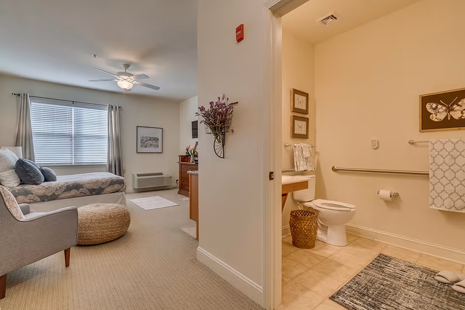 A bedroom suite with a bed, armchair and pouf, and an open doorway to a bathroom showing a toilet, sink and grab bars.