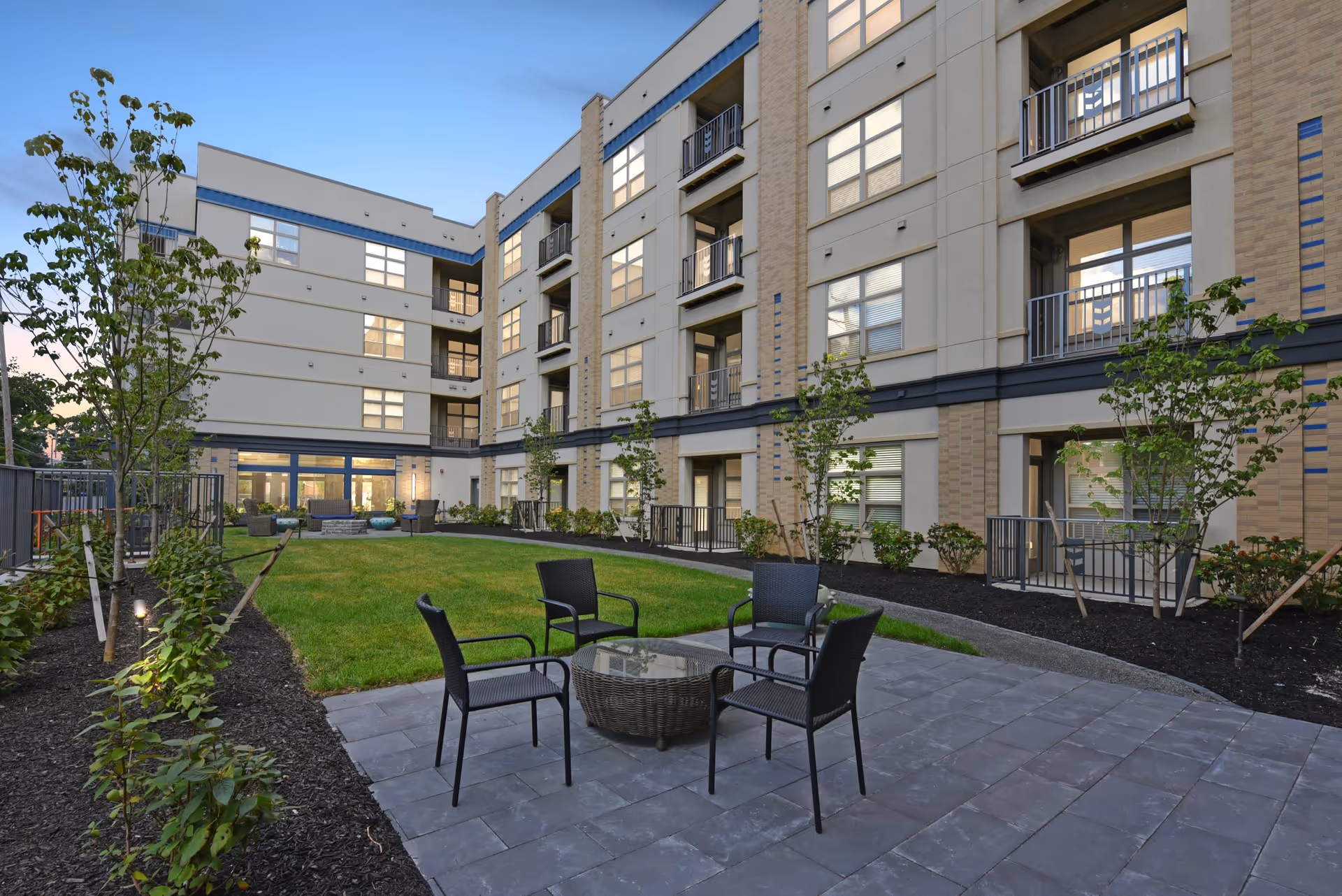 Outdoor courtyard area of a senior living facility named The Deco, featuring a paved patio with four black chairs around a round glass-top table, surrounded by green grass, young trees, and shrubs. The multi-story building with balconies and large windows encloses the courtyard.