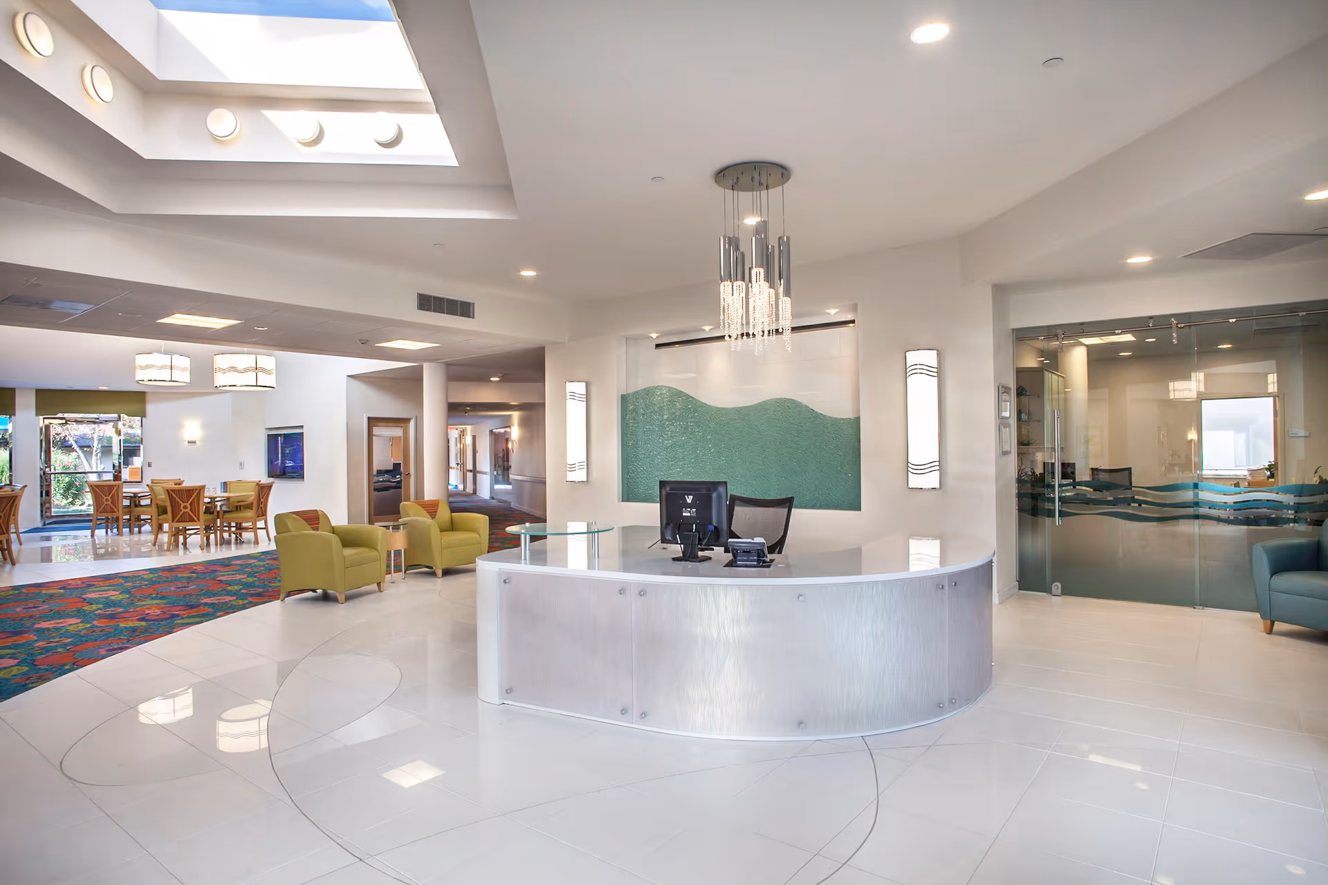 Bright and modern reception area with a curved white desk, computer, and chair. Behind the desk is a decorative wall panel with a wave design and two vertical wall lights. To the left, there are green armchairs and a dining area with wooden tables and chairs. The ceiling has a skylight and contemporary light fixtures. The floor is white tile with circular patterns.