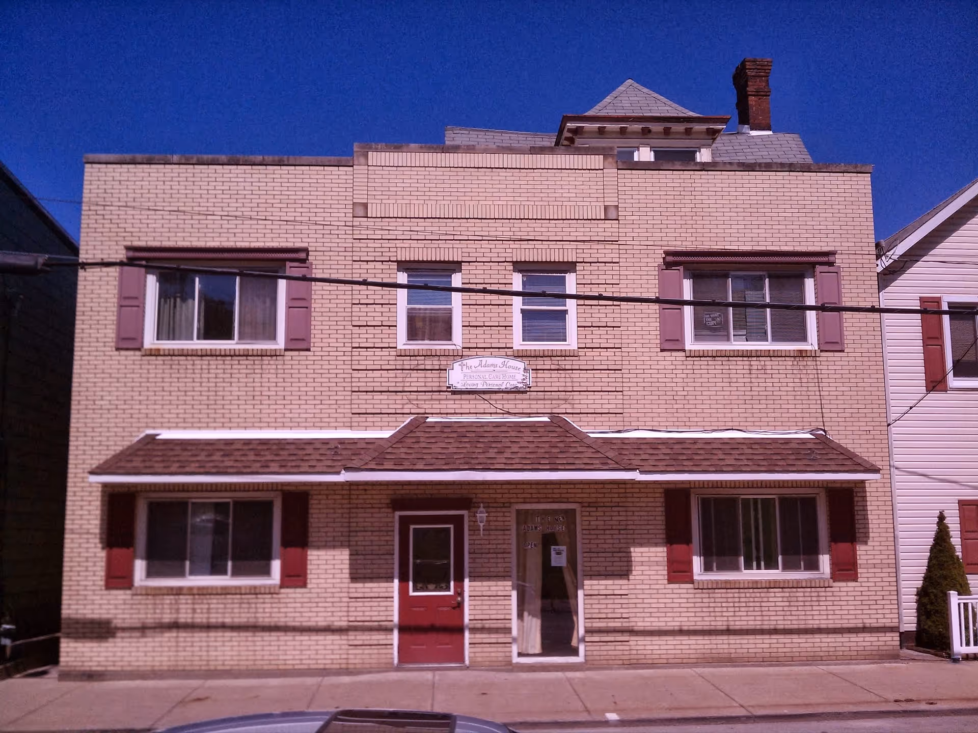 Front exterior view of a two-story brick building with maroon shutters and a small awning above the entrance doors. A sign above the awning reads 'The Adam's House Personal Care Home'. The building is situated on a paved sidewalk under a clear blue sky.