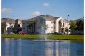 View of a multi-story residential building named Windsor Park Apartments with a pond and a water fountain in the foreground, surrounded by trees and parked cars under a blue sky.
