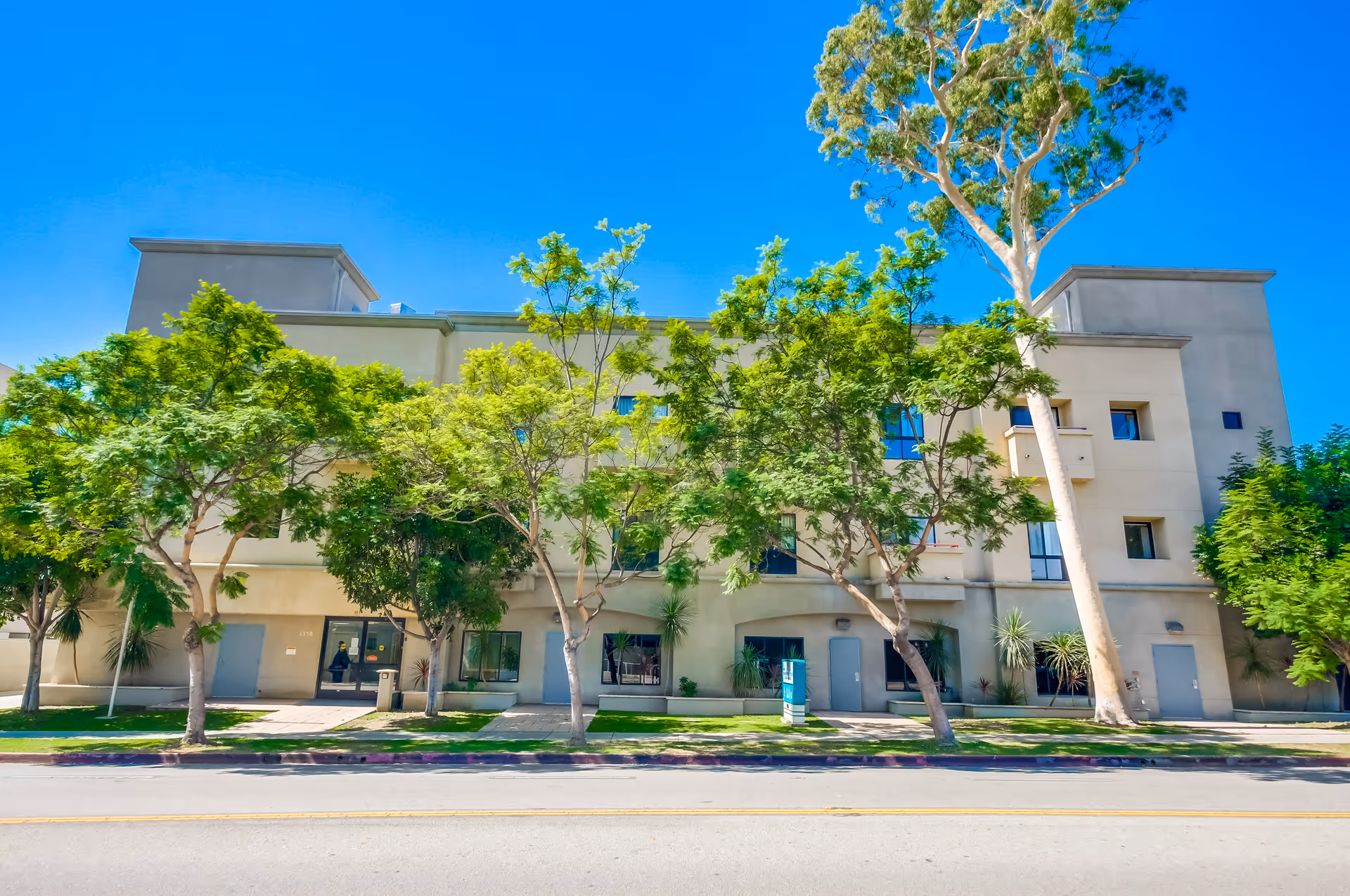 Exterior view of a three-story beige building partially obscured by several green trees under a clear blue sky, with a sidewalk and street in the foreground.