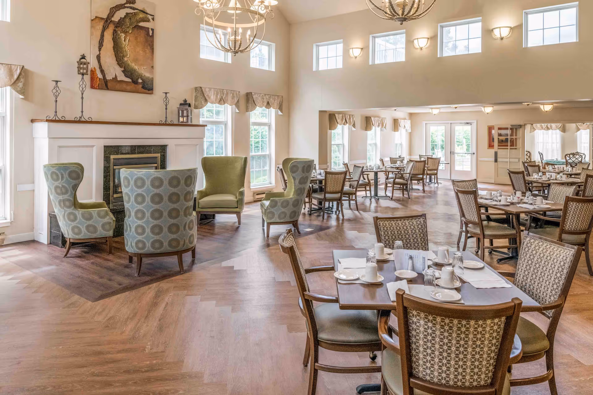 A spacious dining area with multiple tables set with plates, cups, and utensils. The room features high ceilings with several windows allowing natural light to fill the space. There are comfortable armchairs arranged around a fireplace on the left side, and chandeliers hanging from the ceiling. The flooring is a mix of wood patterns, and the walls are painted in a light color.