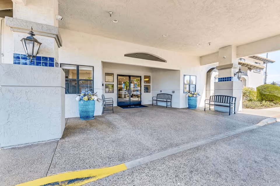 Covered entrance area of Solstice Senior Living at El Cajon with benches, large blue flower pots with floral arrangements, and double glass doors leading inside.