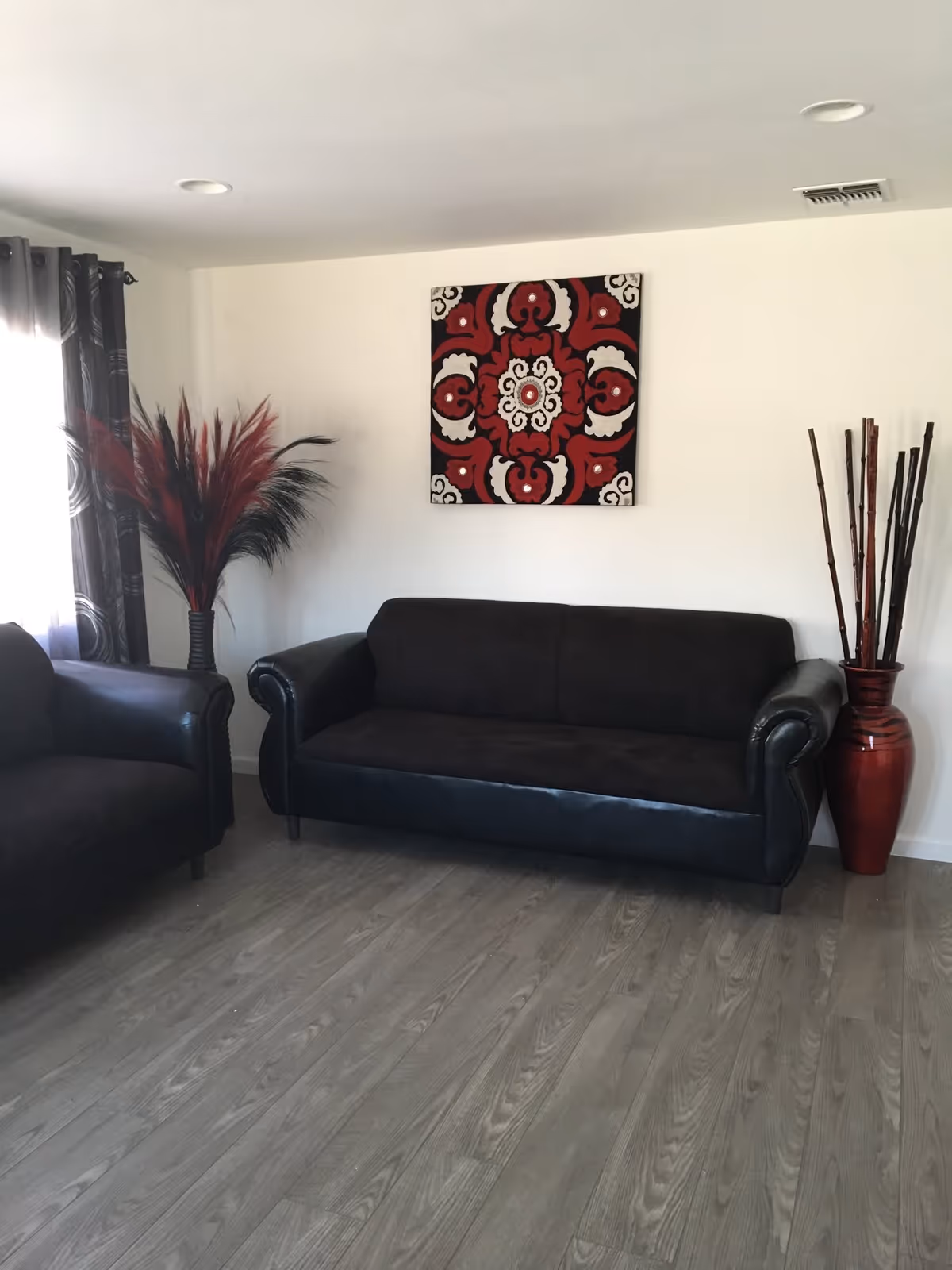 A living room with two black sofas, one on the left and one in the center against a white wall. Above the center sofa is a decorative wall art piece with a red, black, and white pattern. To the left of the sofa is a vase with red and black decorative feathers, and to the right is a tall red vase with long bamboo sticks. The floor is a light wood laminate, and there is a window with dark curtains on the left side.