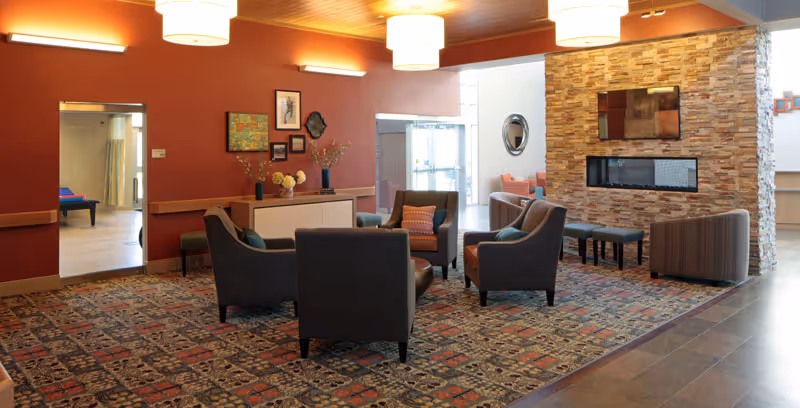 A cozy common area in a senior living facility with a patterned carpet, several armchairs arranged around a small round table, a stone fireplace with a mounted TV above it, and warm lighting. The walls are painted a warm reddish color, and there is a sideboard with decorative items and framed artwork. A doorway leads to another room with exercise equipment visible.