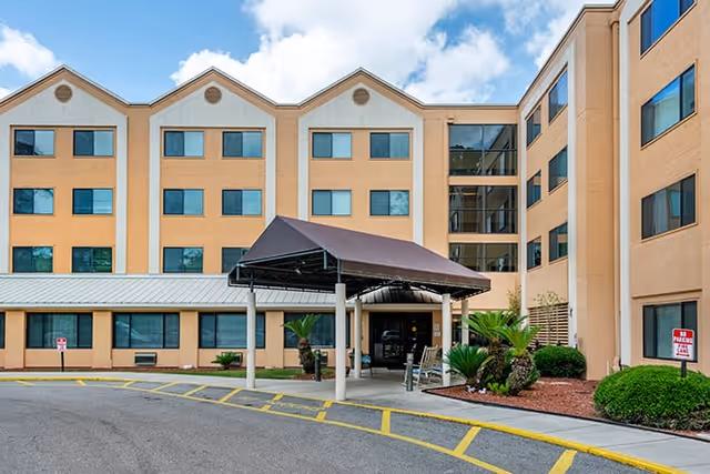Exterior view of a multi-story senior living facility building with a covered entrance, landscaped bushes, and a driveway with yellow painted lines under a partly cloudy sky.