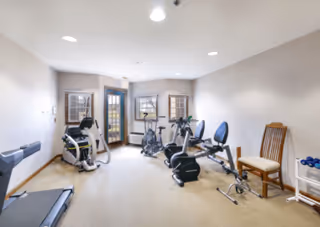 Small indoor fitness room with a treadmill, stationary bikes, weight bench, chair and dumbbells.