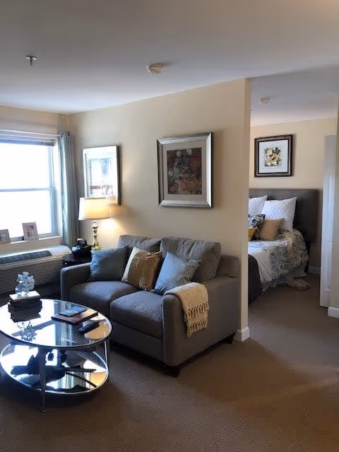 A cozy living area with a gray sofa adorned with pillows and a throw blanket. A round glass coffee table sits in front of the sofa with books and decorative items on it. A lamp on a side table next to the sofa provides warm lighting. The room has beige walls with framed artwork hanging. In the background, a bedroom is partially visible with a bed made up with pillows and a comforter, and framed artwork above the headboard. A window with curtains allows natural light into the living area.