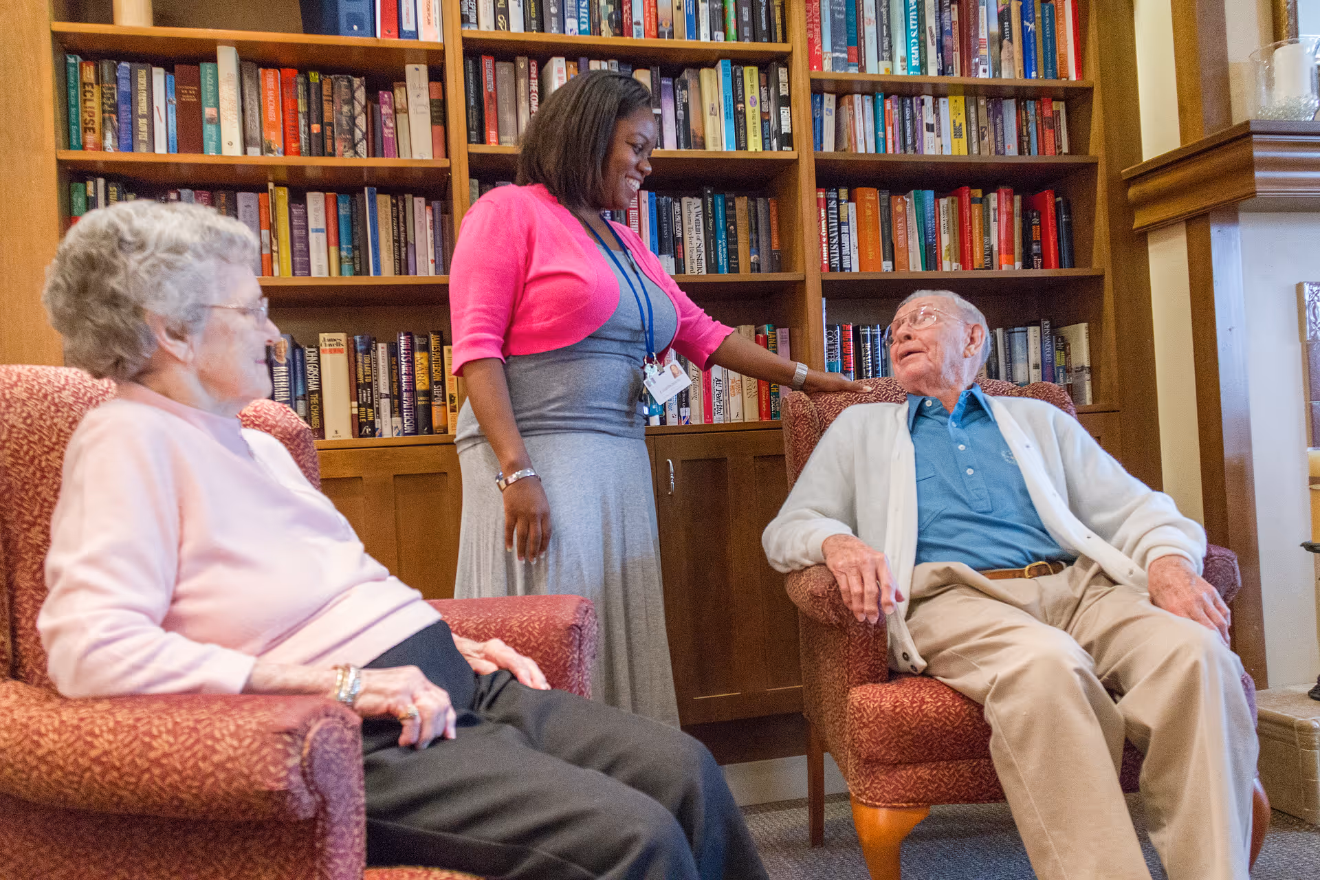 A woman in a pink cardigan and gray dress stands smiling and talking to an elderly man seated in a red armchair. Another elderly woman sits in a similar red armchair nearby. Behind them is a wooden bookshelf filled with books.