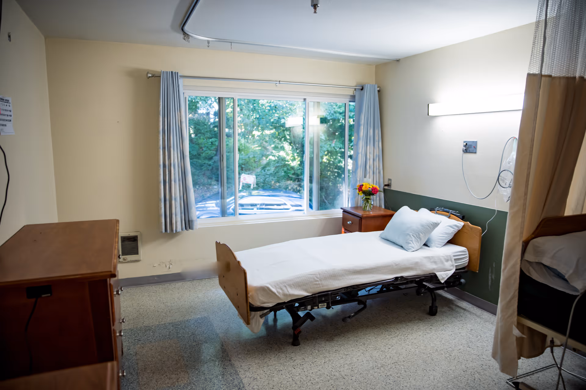 A simple healthcare facility bedroom with a single hospital bed made up with white sheets and pillows, a wooden nightstand with a vase of flowers, a large window with curtains showing greenery outside, and a wooden dresser on the left side.