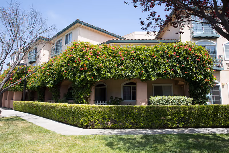 Exterior view of a senior living facility building with beige walls and green roof tiles. The building is partially covered with lush green vines and red flowers. There is a neatly trimmed hedge along the sidewalk and a grassy area in front. Trees with bare branches and some with leaves are visible around the building under a clear blue sky.