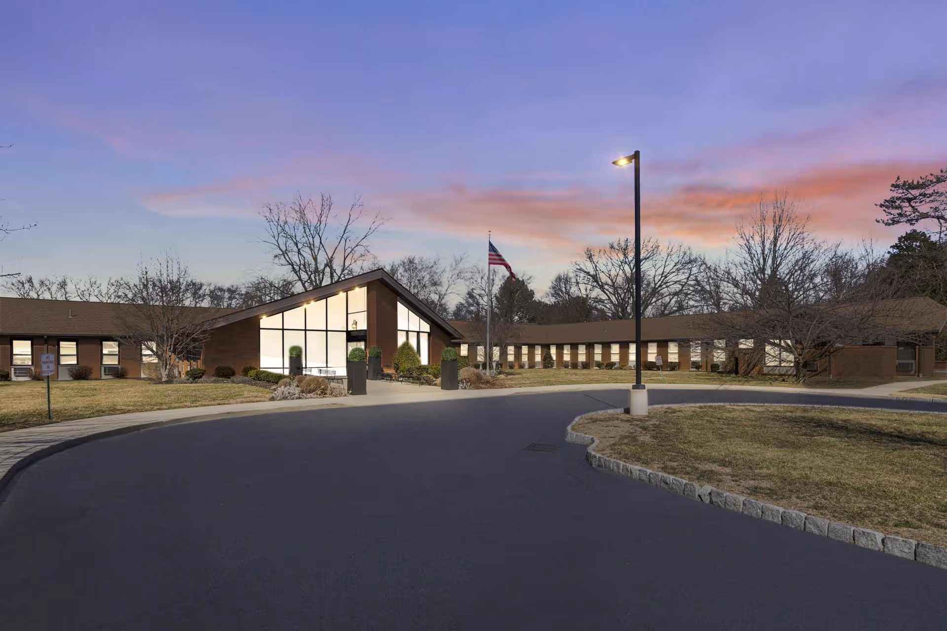 Front exterior of a single-story nursing center with a circular driveway, flagpole and lit entrance at dusk.