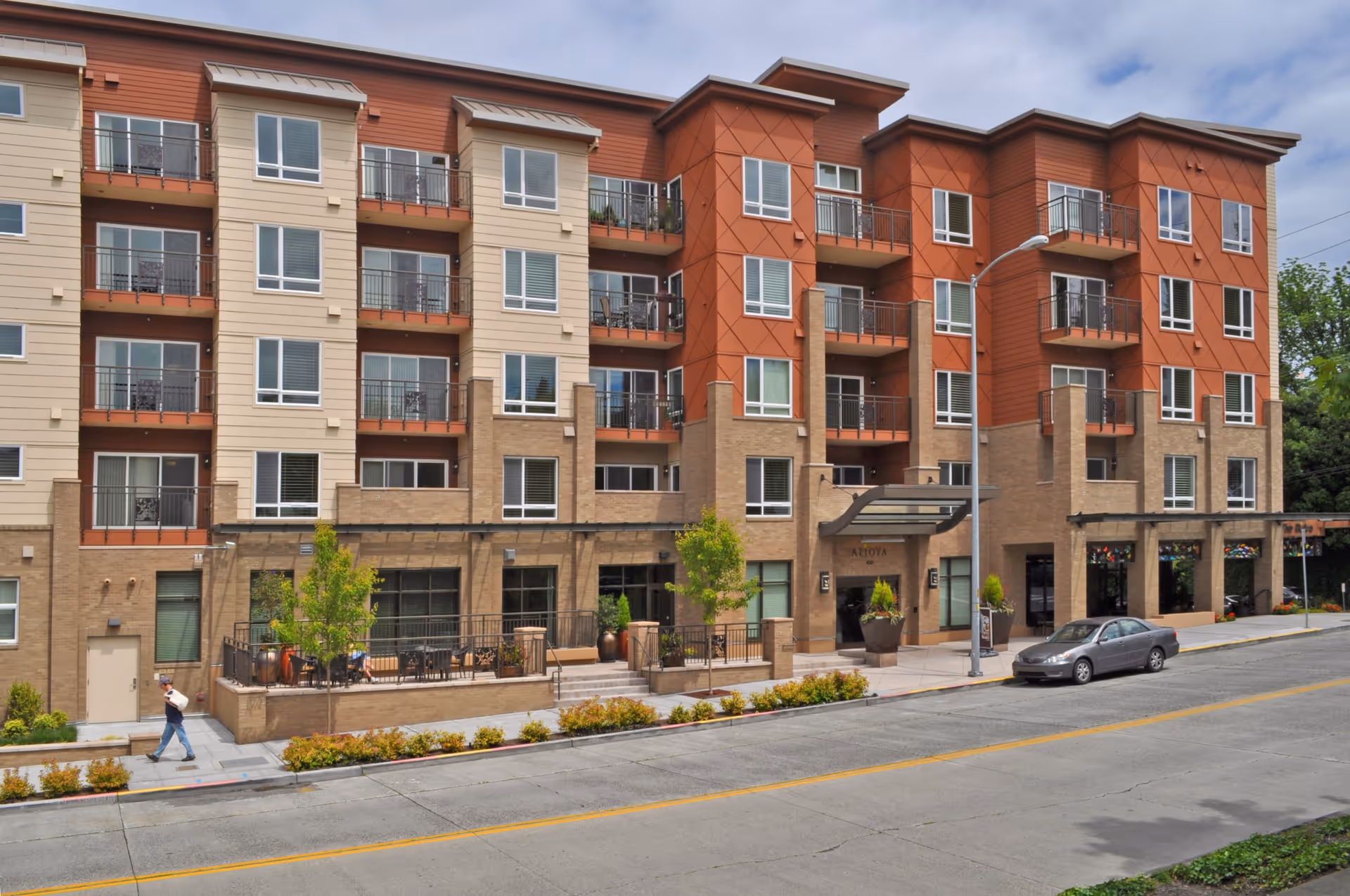 Exterior view of a multi-story senior living facility named Aljoya Thornton Place with balconies, large windows, a covered entrance, a sidewalk, a parked car, and a person walking on the sidewalk.