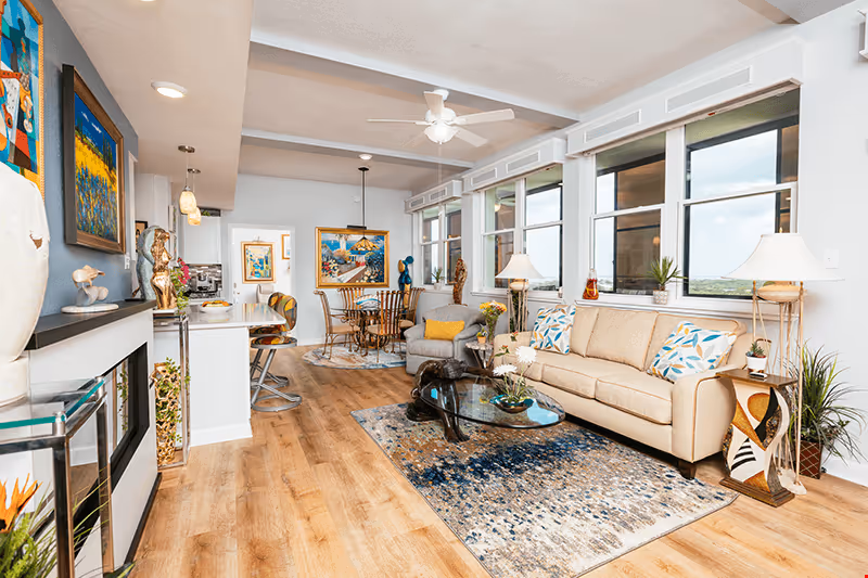 Bright and spacious living room area with a beige sofa adorned with patterned pillows, a glass coffee table on a decorative rug, and a comfortable armchair. The room features large windows letting in natural light, wooden flooring, and various pieces of artwork and sculptures decorating the space. In the background, there is a dining area with a round table and chairs, and a kitchen counter with bar stools.