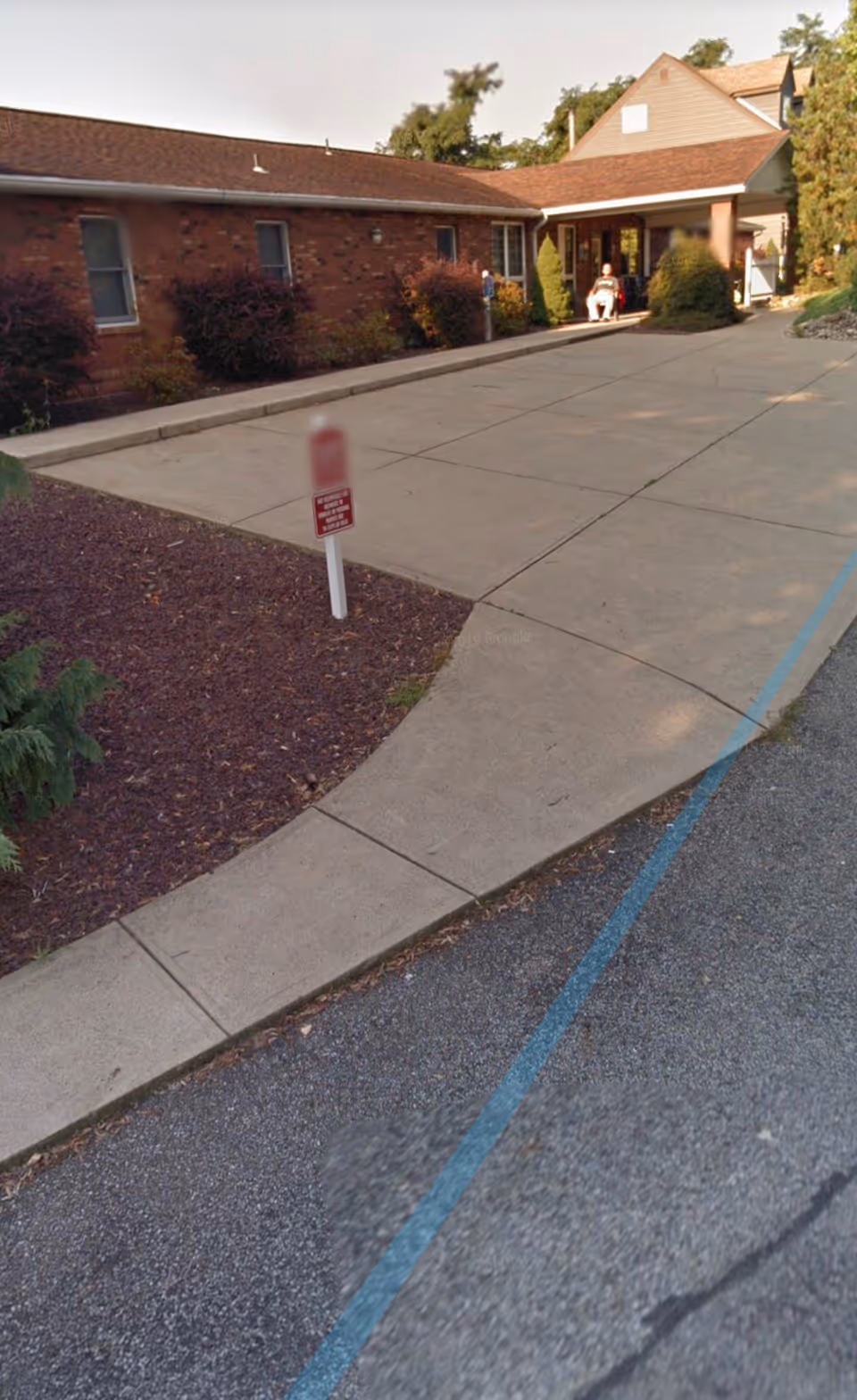 Exterior view of a single-story brick building with a covered entrance. There is a concrete driveway and sidewalk leading to the entrance, with some bushes and landscaping along the side of the building. A person is sitting near the entrance under the covered area.