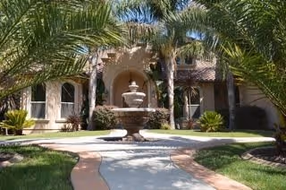 A courtyard entrance of a care home featuring a central stone fountain surrounded by palm trees and a curved walkway leading to the building's arched doorway.